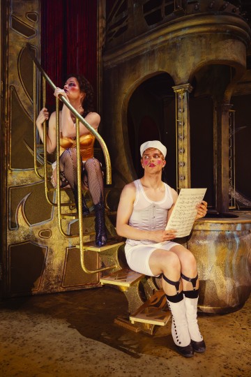 Kaksi dramaattisiin pukuihin ja meikkiin pukeutunutta teatteritaiteilijaa poseeraa kullanvärisessä lavasteessa. Toinen istuu portailla kaiteesta kiinni pitäen, kun taas toinen istuu alhaalla nuotit kädessään ja katsoo eteenpäin.