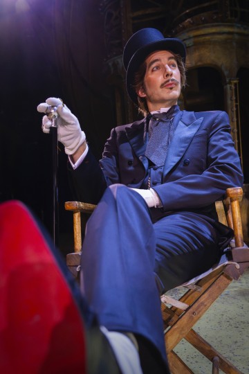 Tyylikkääseen smokkiin, silinterihattuun ja valkoisiin hanskoihin pukeutunut mies istuu itsevarmasti puisella tuolilla, pitää keppiä kädessään ja katsoo ylöspäin hymyillen, ja teatterivalaistus korostaa hänen kasvojaan.