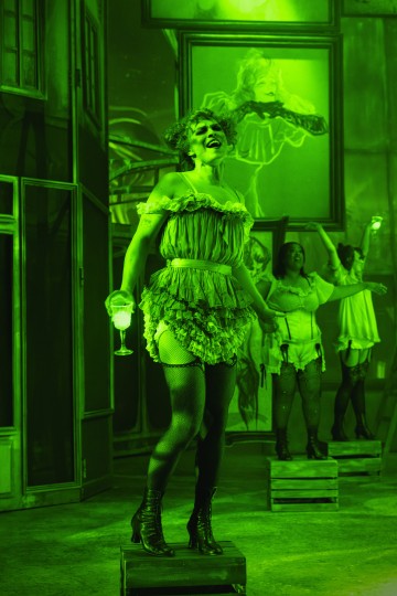 Röyhelöasuun ja verkkosukkiin pukeutunut esiintyjä seisoo laatikon päällä viinilasi kädessään vihreässä valossa kylpevällä näyttämöllä, ja taustalla on kaksi muuta esiintyjää ja kehystettyjä taideteoksia.