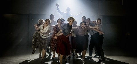 Ryhmä vintage-vaatteisiin pukeutuneita tanssijoita esiintyy tarmokkaasti dramaattisen valaistuksen alla, ja yksi henkilö takana nostaa käsiään, kun valo loistaa hänen takanaan, mikä luo dynaamisen ja teatraalisen kohtauksen.