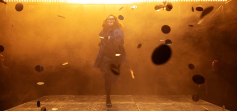 Tummiin vaatteisiin pukeutunut henkilö seisoo lavalla lentävien kolikoiden tai kiekkojen ympäröimänä, ja dramaattinen oranssi valaistus ja sumu luovat dynaamisen, teatraalisen tunnelman.