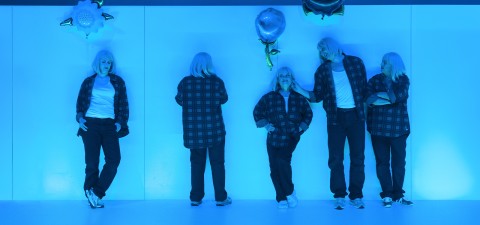 Viisi ihmistä, joilla on lyhyet valkoiset hiukset, mustat ruudulliset paidat, valkoiset T-paidat ja mustat housut, seisovat sinisellä valaistulla seinällä. Joillakin on ilmapalloja ja he ovat vuorovaikutuksessa toistensa kanssa; yksi katsoo seinään päin, toiset eteenpäin tai toisiinsa päin.