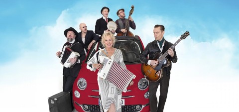 Kuusi muusikkoa seisoo punaisen auton edessä sinistä taivasta vasten. He soittavat erilaisia soittimia, kuten harmonikkaa, banjoa, bassoa ja kitaraa. Edessä oleva nainen hymyilee ja pitää kädessään harmonikkaa.