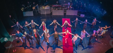 Ryhmä esiintyjiä seisoo lavalla kädet ojennettuina yleisöön päin värikkäiden lavavalojen alla, kun muusikot soittavat soittimia taustalla.