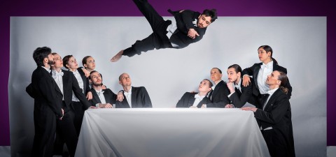 Kaksitoista mustiin pukuihin pukeutunutta ihmistä istuu ja seisoo pöydän ympärillä ja katselee toista henkilöä, joka on heidän yläpuolellaan ilmassa, ikään kuin hyppäämässä tai kääntymässä, tavallista taustaa vasten.