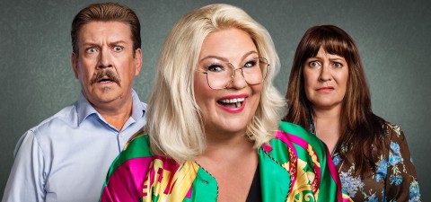 Kolme aikuista seisoo harmaata taustaa vasten. Edessä hymyilevä nainen, jolla on vaaleat hiukset ja suuret silmälasit, on pukeutunut värikkääseen paitaan. Hänen takanaan viiksekäs mies ja ruskeatukkainen nainen näyttävät molemmat yllättyneiltä tai huolestuneilta.