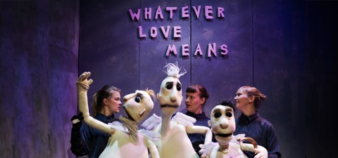 Neljä tummiin vaatteisiin pukeutunutta nukketeatteritaiteilijaa käyttää suuria valkoisia nukkeja seinän edessä, jossa on violeteilla kirjaimilla teksti WHATEVER LOVE MEANS. Kohtaus on valaistu dramaattisella violetilla valaistuksella.