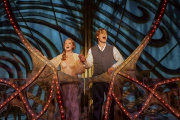 Persikkaiseen mekkoon pukeutunut nainen ja liiviin ja paitaan pukeutunut mies seisovat kädestä pitäen ja laulavat iloisesti kirkkaasti valaistulla, koristeellisella näyttämöllä, jonka lavasteissa on pyöriviä kuvioita ja punaisia valoja.