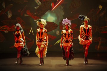 Neljä naista kävelee lavalle itsevarmasti yllään oranssin ja mustan korsetin puvut, höyhenpäähineet ja korkeat saappaat, ja heidän takanaan on dramaattinen, värikäs tausta.