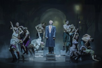 Siniseen takkiin pukeutunut mies seisoo keskellä näyttämöä laatikolla, jota ympäröivät dramaattisissa, ilmeikkäissä asennoissa ja teatteripuvuissa olevat tanssijat, tunnelmallisessa valaistuksessa ja sumuisessa, tunnelimaista taustaa vasten.