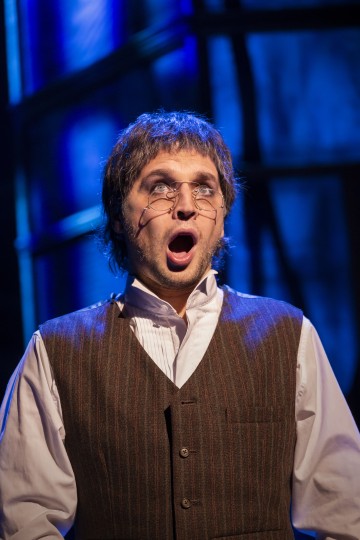 Silmälasit ja raidallinen liivi päällään oleva henkilö seisoo suu auki ja vaikuttaa yllättyneeltä tai järkyttyneeltä dramaattisessa sinisessä näyttämövalaistuksessa.