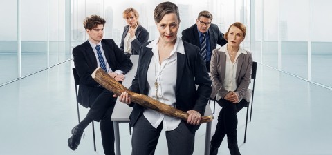 Viisi vakavasti otettavaa pukumiestä istuu ja seisoo modernissa, valoisassa toimistossa. Keskellä seisova nainen pitää kädessään suurta puista mailaa, ja hänen takanaan istuu neljä kollegaa, jotka näyttävät päättäväisiltä.