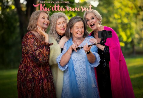 Neljä vanhempaa naista ottaa hymyillen ja poseeraten yhdessä ulkona selfien älypuhelimella. He ovat pukeutuneet värikkäisiin, tyylikkäisiin vaatteisiin. Auringonvalo suodattuu taustalla olevien vihreiden puiden läpi. Kuvan yläreunassa näkyy teksti.