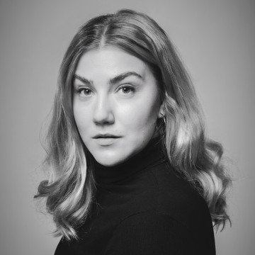 Mustavalkoinen muotokuva naisesta, jolla on pitkät, aaltoilevat hiukset ja musta poolopaita. Hän katsoo suoraan kameraan neutraalilla ilmeellä tavallista taustaa vasten.