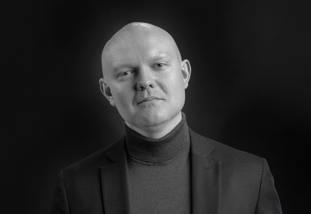 Mustavalkoisessa muotokuvassa henkilö, jolla on ajeltu pää ja jolla on tumma poolokaulus ja bleiseri, katsoo suoraan kameraan tummaa taustaa vasten.