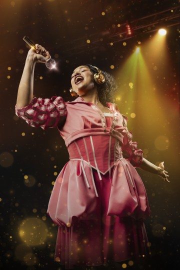 Vaaleanpunaiseen mekkoon ja kultaisiin kuulokkeisiin pukeutunut nainen laulaa intohimoisesti lavalla kimaltelevaan mikrofoniin, ja taustalla on värikkäitä valonheittimiä ja kultaisia bokeh-valoja.