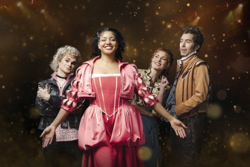 Neljä teatteripukuista näyttelijää seisoo yhdessä hymyillen. Keskushahmolla on kirkkaan vaaleanpunainen, taidokas mekko, muilla on yllään eklektiset asusteet. Taustalla on hehkuva, kultainen bokeh-efekti, joka luo juhlatunnelmaa.