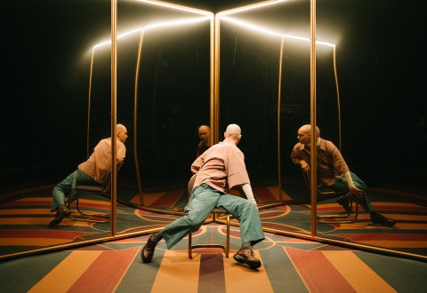 Henkilö satunnaisesti pukeutuneena istuu tuolilla suurten peilien edessä, mikä luo useita heijastuksia. Tilassa on dramaattinen valaistus ja värikäs, geometrinen lattiakuvio.