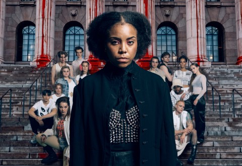 Tummiin vaatteisiin pukeutunut nainen seisoo itsevarmasti vakavasti otettavien ihmisten ryhmän edessä kiviportailla, joiden takana olevissa pylväissä on punaisia raitoja. Rakennus on mahtipontinen suurine kaari-ikkunoineen.