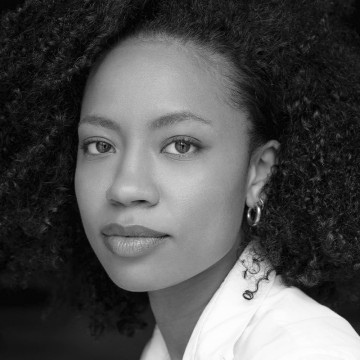 Mustavalkoinen muotokuva kiharatukkaisesta naisesta, jolla on korvakorut ja vaalea toppi ja joka katsoo suoraan kameraan rauhallinen ilme kasvoillaan.