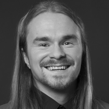 Pitkätukkainen ja parrakas mies hymyilee kameralle. Hänellä on yllään tumma paita, ja tausta on tavallinen ja tumma. Kuva on mustavalkoinen.