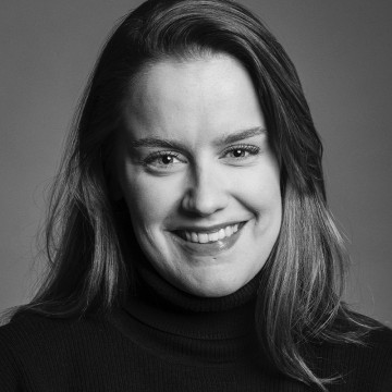 Mustavalkoinen muotokuva naisesta, jolla on pitkät hiukset ja joka hymyilee hellästi. Hänellä on yllään tumma poolopusero ja hän katsoo suoraan kameraan tavallista taustaa vasten.