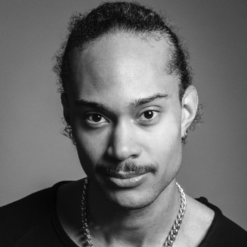 Mustavalkoinen muotokuva henkilöstä, jolla on taakse vedetyt kiharat hiukset ja viikset. Hänellä on kaulakoru, korvakorut ja tumma paita, ja hän katsoo suoraan kameraan neutraali ilme kasvoillaan.