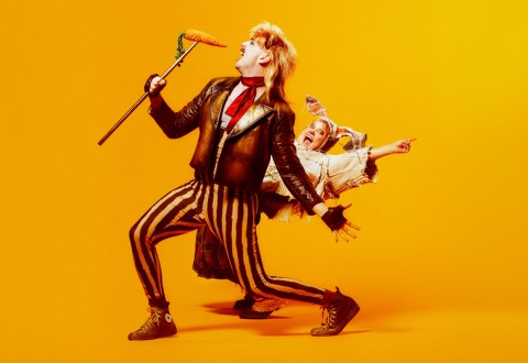 Kaksi teatteripukuista ihmistä poseeraa dramaattisesti kirkkaan keltaista taustaa vasten. Toisella on nahkatakki ja raidalliset housut, ja hänellä on kädessään maissimaisemainen valtikka; toinen, riekaleisiin vaatteisiin pukeutunut, nojaa taaksepäin ja osoittaa innoissaan.